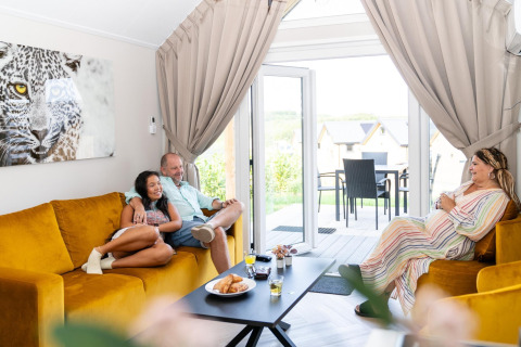 Tre personer hygger sig i en stue med gule sofaer på Berg Lodge i Holland, sollys strømmer ind fra terrassen.