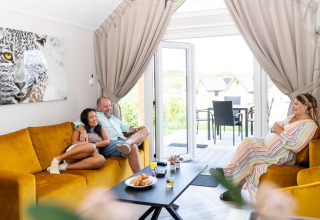 Tre personer hygger sig i en stue med gule sofaer på Berg Lodge i Holland, sollys strømmer ind fra terrassen.