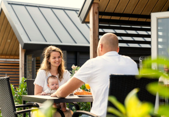 Une famille avec un bébé prend un repas en extérieur à Berg Lodge aux Pays-Bas, lodges en bois en arrière-plan.