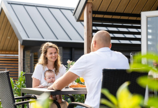 En familie med baby nyder et måltid udenfor på Berg Lodge i Holland, moderne træhytter i baggrunden.