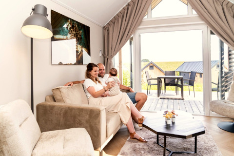 Gezin ontspant zich in de lichte woonkamer van Berg Lodge in Nederland, uitzicht op terras en huisjes.