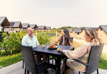 Three people enjoy a meal and conversation outside Berg Lodge in the Netherlands with modern lodges nearby.