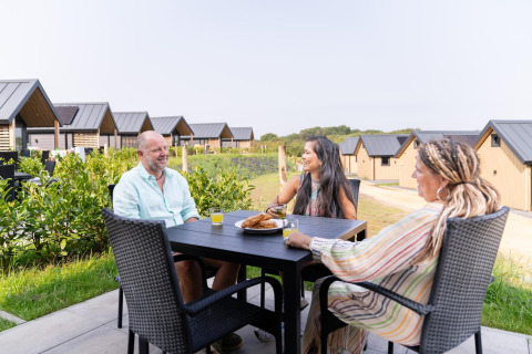 Tre personer hygger sig udendørs ved et bord foran Berg Lodge i Holland, omgivet af moderne hytter.