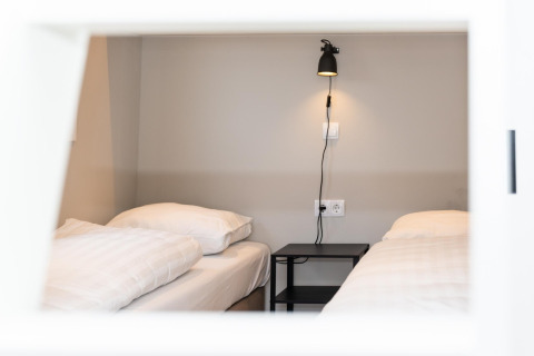 To enkeltsenge med hvidt sengetøj og et sort natbord på Berg Lodge i Holland, set gennem et vindue.