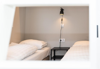 To enkeltsenge med hvidt sengetøj og et sort natbord på Berg Lodge i Holland, set gennem et vindue.