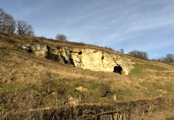 View of a hillside with a rocky cave entrance and sparse grass beneath a bright blue sky.