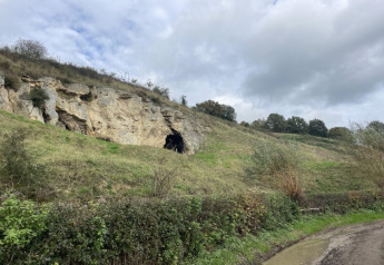 Photo of a steep hillside with a cave entrance, green vegetation and cloudy sky at Berg Lodge, Netherlands.