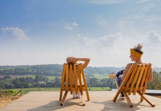 Deux personnes se détendent sur des chaises en bois, profitant d’une vue sur la campagne depuis la terrasse d’un lodge.