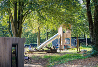 Des enfants jouent sur une aire de jeux en bois avec toboggan près d’un lodge entouré d’arbres.