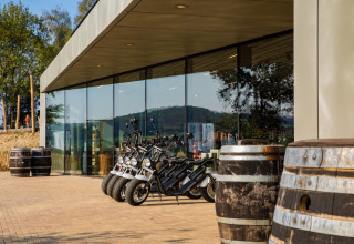 Lodge moderne avec murs en verre, scooters électriques garés et grands tonneaux en bois à l’extérieur.