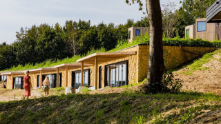 Two people walk past hillside lodges with green roofs at a lodge, surrounded by trees in summertime.