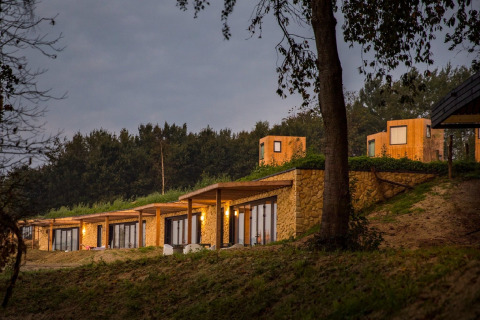 Photo de lodges chaleureux aux toits végétalisés et murs en pierre, nichés au bord d’une forêt au crépuscule.