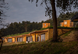 Photo de lodges chaleureux aux toits végétalisés et murs en pierre, nichés au bord d’une forêt au crépuscule.