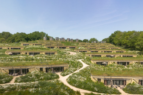 Lodges écologiques intégrés dans la colline, toits verts, fleurs sauvages, sentiers et arbres environnants.