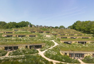 Lodges écologiques intégrés dans la colline, toits verts, fleurs sauvages, sentiers et arbres environnants.