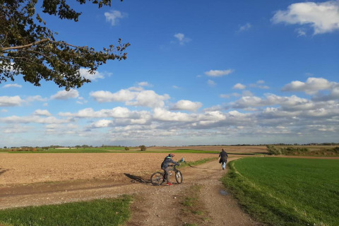 Personne avec un vélo et une autre marchant sur un chemin de campagne sous un ciel bleu parsemé de nuages.