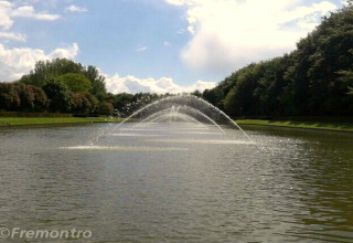 Un bassin paisible avec une grande fontaine au centre, entouré d’arbres et de verdure sous un ciel bleu.