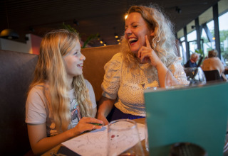 Une mère et sa fille aux longs cheveux blonds rient ensemble à une table d'un restaurant de lodge.
