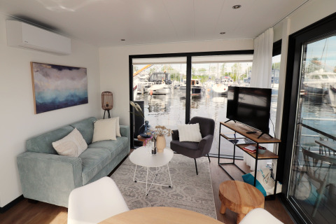 Salon du houseboat 'Refugio' aux Pays-Bas, avec canapé, fauteuil, TV et vue sur le port de plaisance.