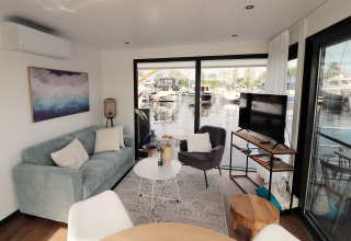 Salon du houseboat 'Refugio' aux Pays-Bas, avec canapé, fauteuil, TV et vue sur le port de plaisance.