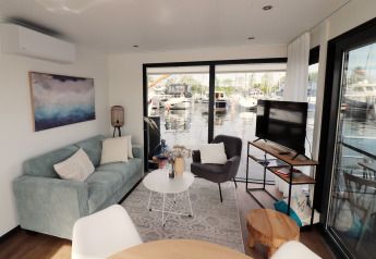 Salon du houseboat 'Refugio' aux Pays-Bas, avec canapé, fauteuil, TV et vue sur le port de plaisance.