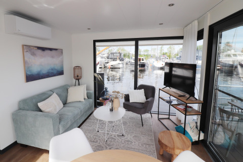 Stue på husbåd 'Refugio' i Holland med sofa, stol, tv og store vinduer med udsigt over en havn.