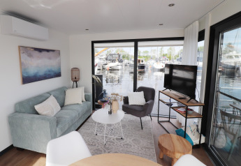 Soggiorno su houseboat 'Refugio' nei Paesi Bassi con divano, poltrona, TV e vista sul porto turistico.