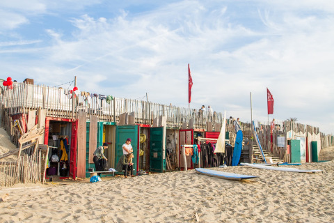 Glampingverblijf op een zandstrand met kleurrijke hutten, surfplanken en mensen die relaxen.