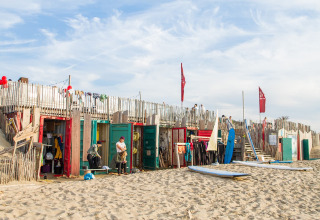 Glampingverblijf op een zandstrand met kleurrijke hutten, surfplanken en mensen die relaxen.