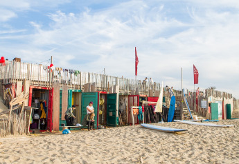 Hébergement glamping sur une plage de sable avec cabanes colorées, planches de surf et vacanciers.
