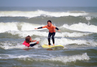 Een kind leert surfen met begeleiding van een instructeur vlakbij een glamping-verblijf aan zee.
