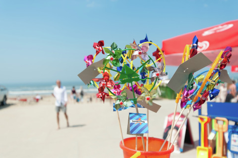 Kleurrijke windmolentjes te koop op het strand bij een glampingverblijf, met zee en parasols op de achtergrond.