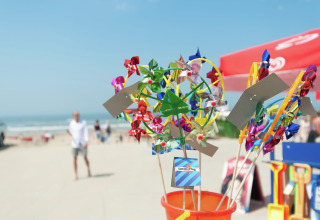 Kleurrijke windmolentjes te koop op het strand bij een glampingverblijf, met zee en parasols op de achtergrond.