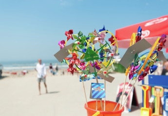Moulins à vent colorés en vente sur la plage d’un site de glamping, avec la mer et des parasols en arrière-plan.