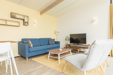 Lichte moderne woonkamer in Sea Lodges Ameland 2 met blauwe zetel, tv en gele accenten in Nederland.