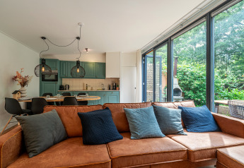 Bright lodge living room with brown leather sofa, colorful pillows, open kitchen, and large windows to garden.