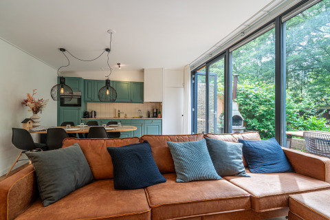 Moderne lodge stue med brun sofa, farverige puder, åbent køkken og store vinduer med udsigt til naturen.