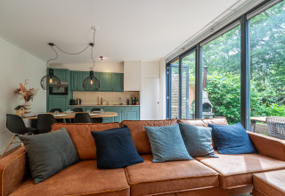 Moderne lodge stue med brun sofa, farverige puder, åbent køkken og store vinduer med udsigt til naturen.