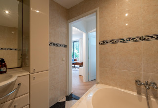 Badeværelse i Wellnes suite-lodge i Holland, med badekar, flisevæg og udsigt til stue via åben dør.
