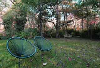 Twee groene stoelen en een houten bankje in een rustige tuin van een lodge, omgeven door bomen.