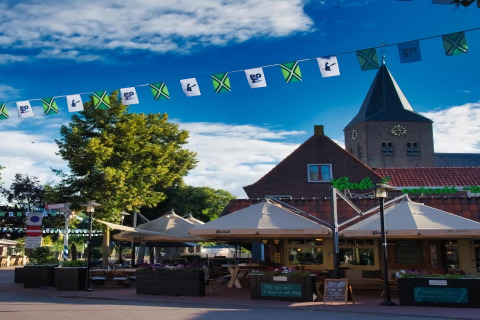 Udendørs café foran et lodge med solbeskinnet terrasse, flag, grønne træer og en kirke i baggrunden.