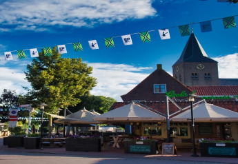 Gezellig terras aan een lodge met vlaggetjes en parasols, bomen en een kerk zichtbaar onder een blauwe lucht.