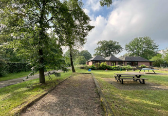 Lodge met picknicktafels, veel groen en een pad onder een deels bewolkte hemel op een zomerse dag.