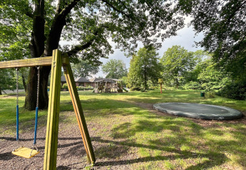 Buitenspeeltuin bij een lodge met schommel, trampoline, speeltoestel en groene bomen rondom.