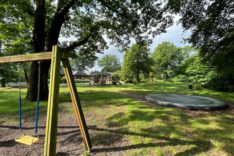 Spielplatz im Freien bei einer Lodge mit Schaukel, Trampolin, Klettergerüst und umgebenden Bäumen.