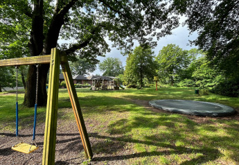 Parque infantil al aire libre en una cabaña con columpio, trampolín, tobogán y árboles verdes alrededor.