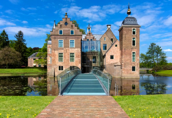 Foto van een historisch landhuis met moderne glazen brug over een gracht, omringd door natuur en blauwe lucht.