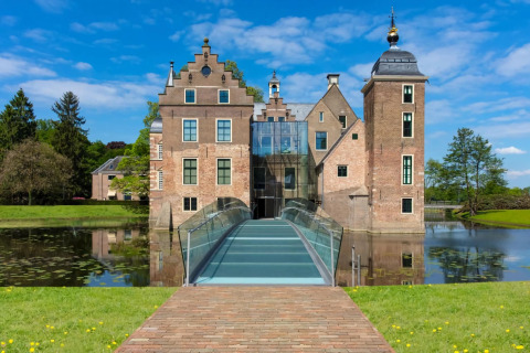 Photo of a historic lodge with a modern glass bridge over a moat, surrounded by greenery and blue sky.