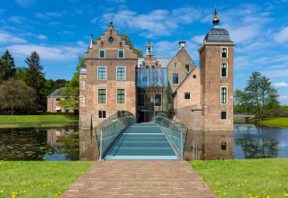Afbeelding van een historische lodge met moderne glazen brug over een gracht, met groen en blauwe lucht.
