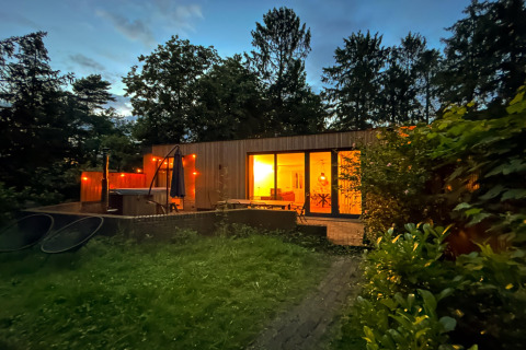Aftenfoto af Wellnes suite, en lodge i Holland, med oplyst interiør og jacuzziterrasse omgivet af skov.